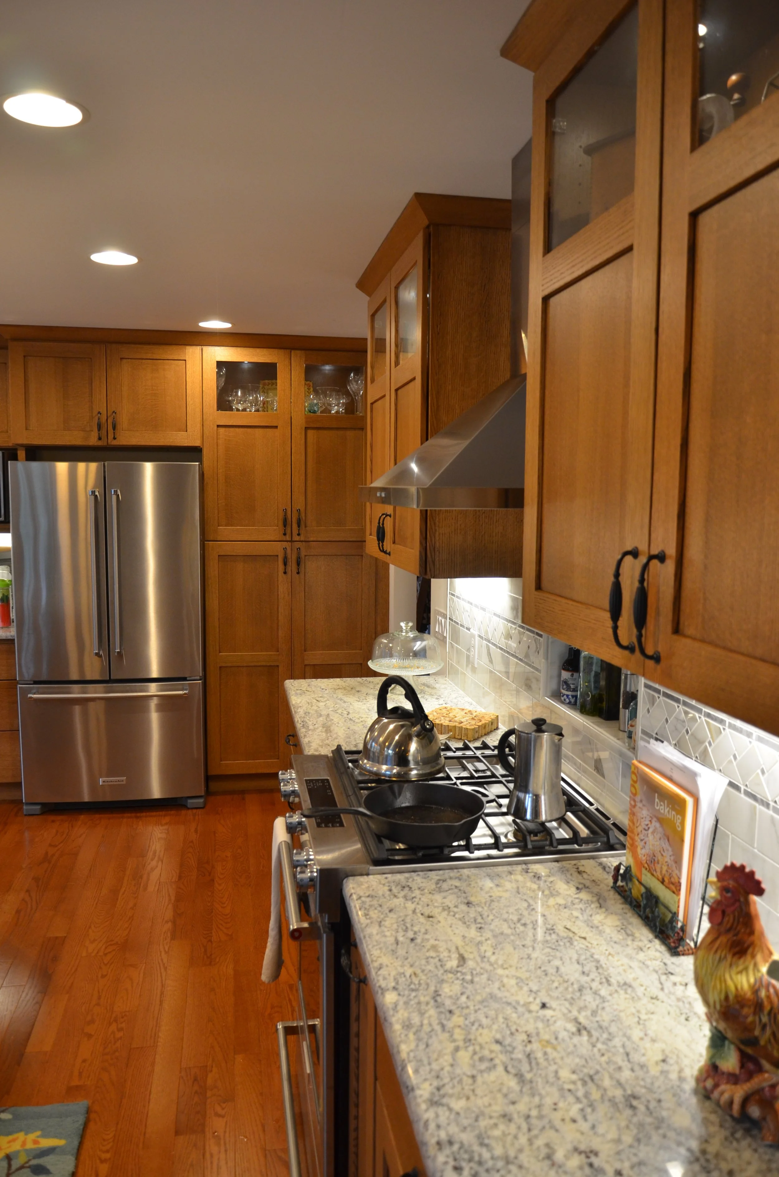 A kitchen with wooden cabinets, a stainless steel refrigerator, a gas stove with a kettle and a pan, a granite countertop, a rooster figurine, and a cookbook or magazine on the counter.