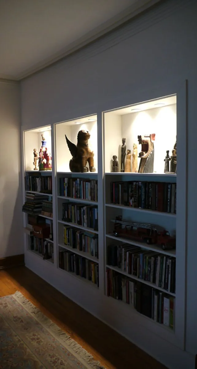 White bookshelf with three lit display cases on top, featuring various sculptures and artifacts, against a wall in a room with wooden floor and a decorative rug.