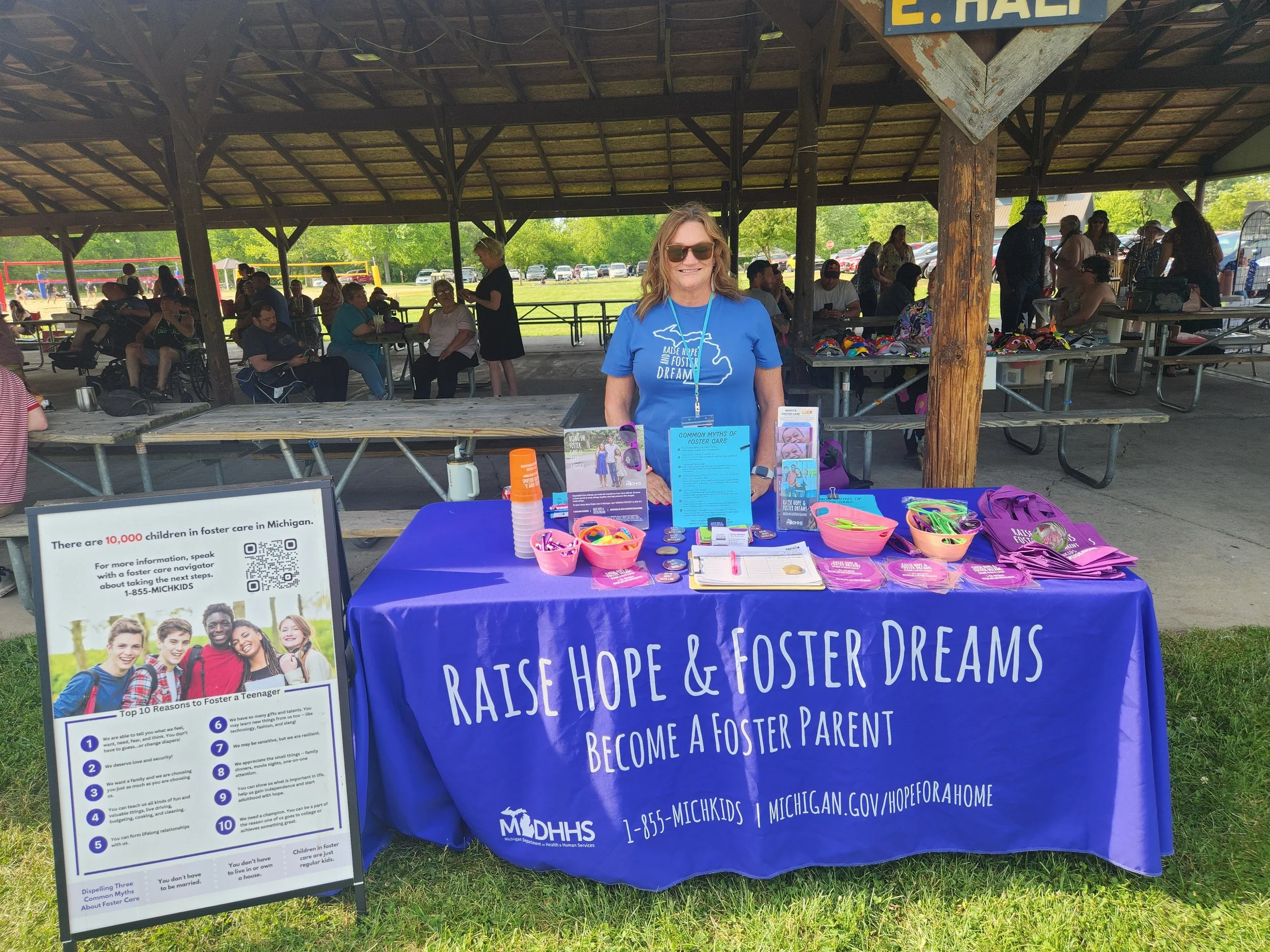 A woman in sunglasses standing behind a table with informational materials at an outdoor event in a pavilion. The table promotes foster care awareness, with purple tote bags and brochures. There are other people in the background sitting and walking 