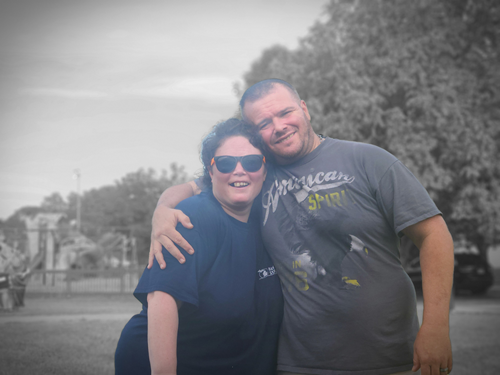A smiling woman wearing sunglasses and a dark blue shirt standing beside a smiling man wearing a gray T-shirt, outdoors with trees and a cloudy sky in the background.