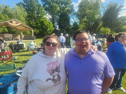 Two people smiling at an outdoor gathering with trees and other attendees in the background.
