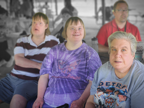 A group of four people, including a young woman with Down syndrome, sitting in a room, with some in the background. They are looking in different directions.