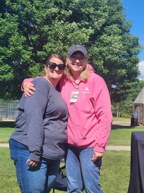 Two women smiling and hugging outdoors in a park on a sunny day, with trees and a playground in the background. One woman wears sunglasses and a gray sweatshirt, the other wears a pink hoodie, a gray cap, and sunglasses.