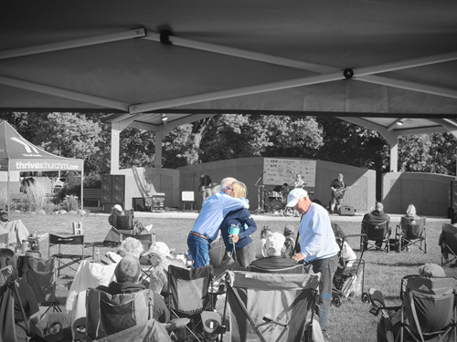 People attending an outdoor event or concert under a tent, with a stage in the background, some seated in chairs, some standing, with volunteers interacting with attendees.