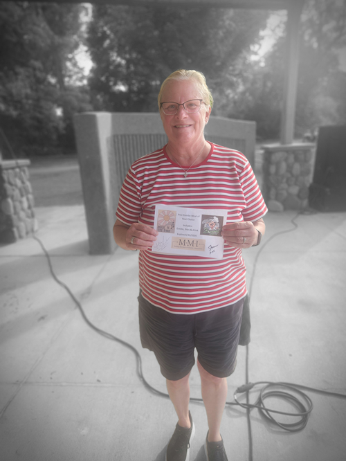 An elderly woman with blonde hair, glasses, wearing a red and white striped shirt and dark shorts, standing outdoors on a concrete pavement, holding a certificate and smiling.