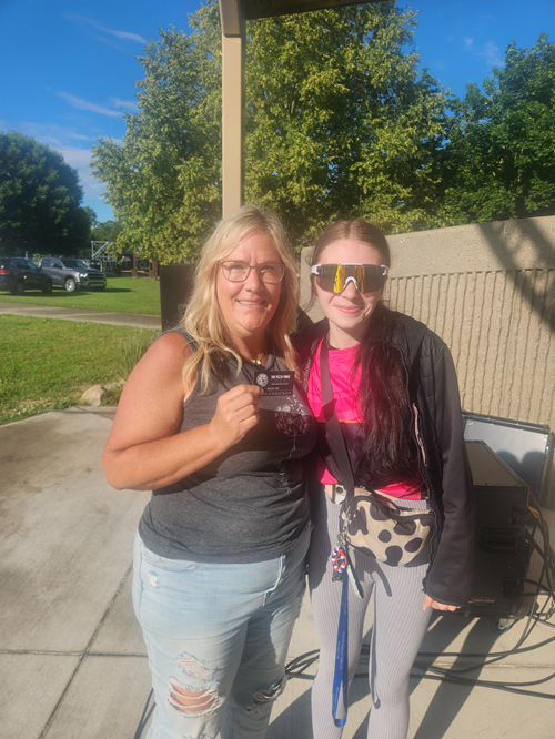 Two women smiling outdoors on a sunny day, one holding a card, the other wearing sunglasses and a small bag, standing near a concrete wall and some equipment.