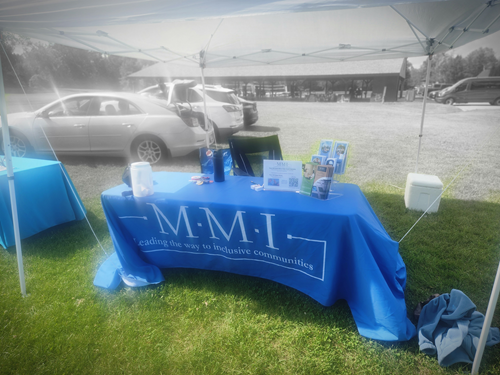 Information table with pamphlets and supplies under a white canopy at an outdoor event, with parked cars and a building in the background.