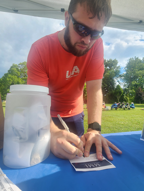 Man with sunglasses wearing a red UA t-shirt, signing a document on a blue table outside under a canopy, with trees and a grassy field in the background.