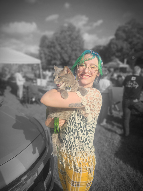 A woman with colorful hair and glasses holding a cat outdoors at a park or event, with other people and trees in the background.