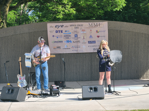Musicians performing on an outdoor stage with a banner in the background, in front of a wooden wall and trees.
