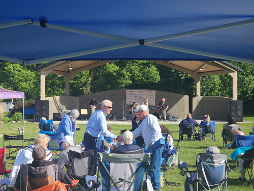 Outdoor community event with people gathered under a gazebo, some seated in chairs, others standing and talking, with a band performing on stage in the background.