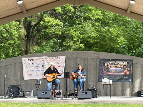 Two musicians playing guitars on an outdoor stage with green trees in the background