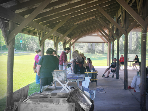 People browsing art tables inside a wooden pavilion at an outdoor park.