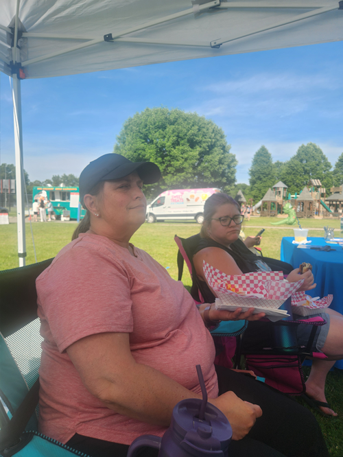 Two women sitting outdoors under a tent at a park or fair, with trees and a playground in the background. One woman is wearing a pink shirt and black cap, and the other is wearing glasses and a black shirt, eating food.