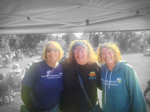 Three smiling women standing under a white canopy at an outdoor event.