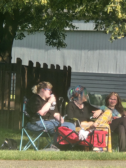 Three women sitting outdoors under a tree, relaxing and chatting. Two women are sitting on camping chairs, and the third is sitting on the grass. They appear to be enjoying warm weather and casual conversation.