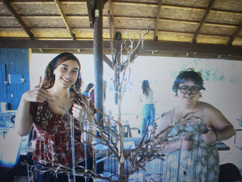 Two women smiling at a craft or art display, with a bare tree branch in front of them at an outdoor setting under a wooden roof.