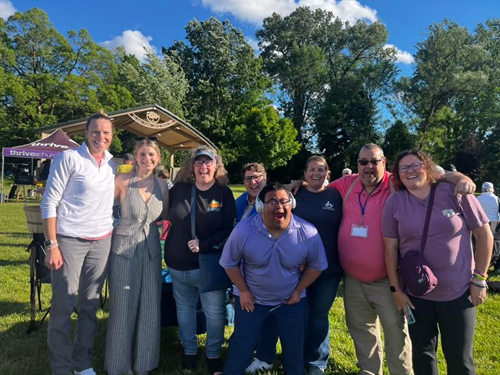 Group of eight people smiling outdoors in a park, some wearing sunglasses, with trees and a blue sky with clouds in the background.