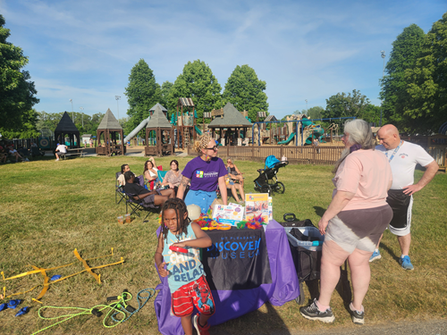 Parents and children at an outdoor community event with a playground in the background, featuring a table with a 'Discover' sign, toys, and informational materials, on a sunny day.