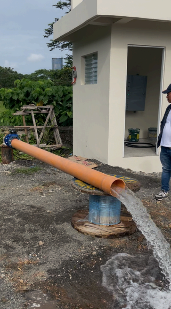 A large orange pipe is releasing water into a makeshift fountain made from a blue plastic barrel on a wooden surface. A man stands nearby, observing the setup outside a small beige building with an open door and electrical panel. There is greenery and construction materials in the background.