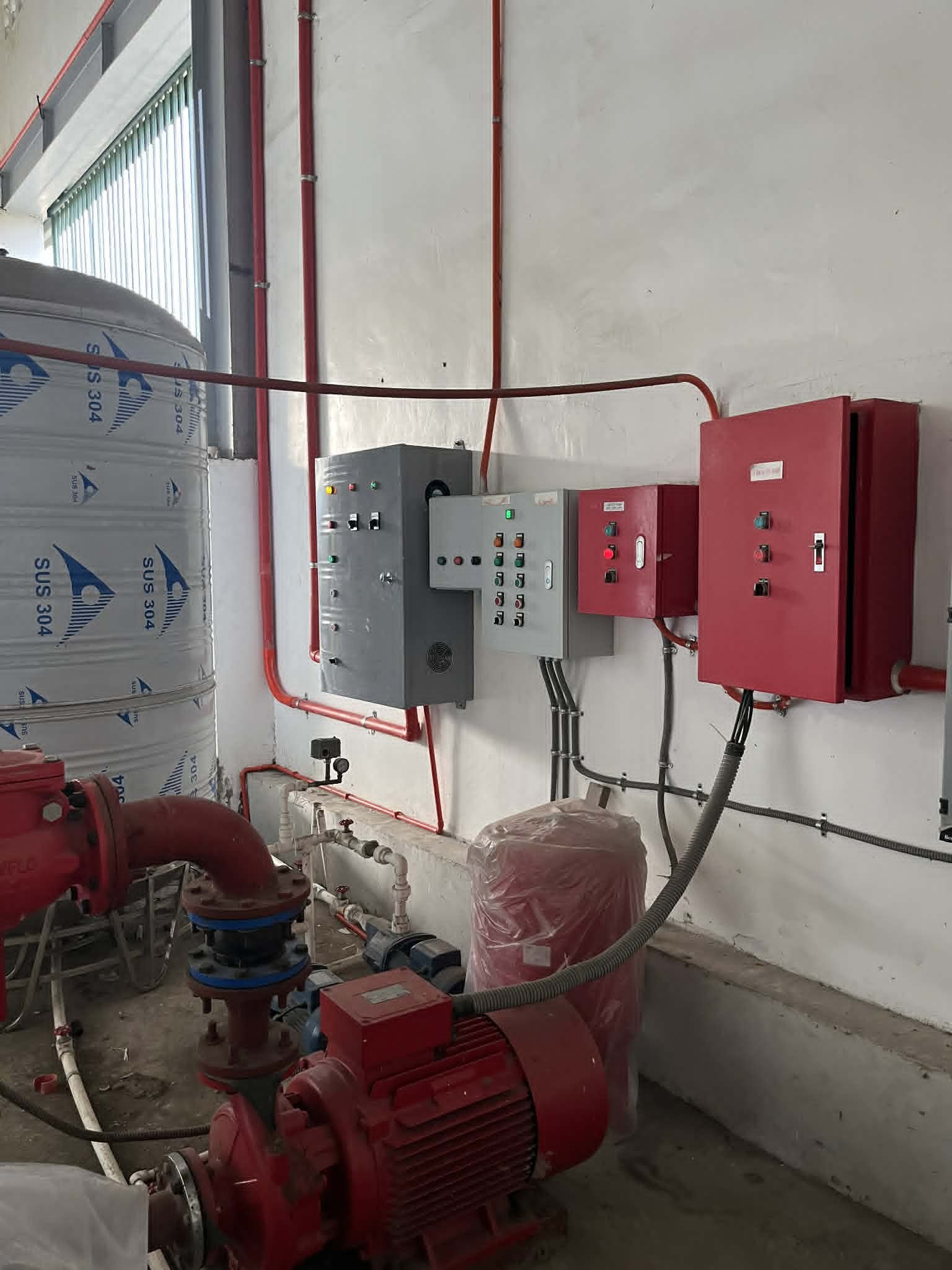 Industrial equipment including a red motor pump, control panels, insulated storage tanks, pipes, and electrical wiring in a utility room.