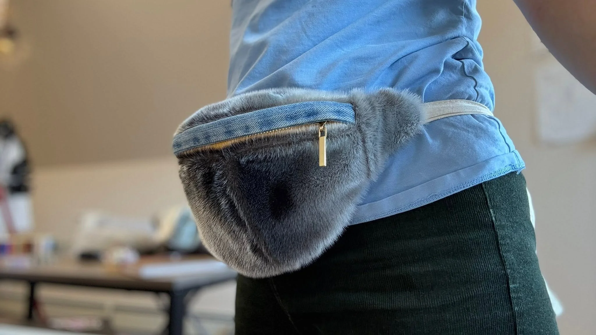 Close-up of a person's side showing their blue shirt, green pants, and a gray furry purse with a gold zipper.