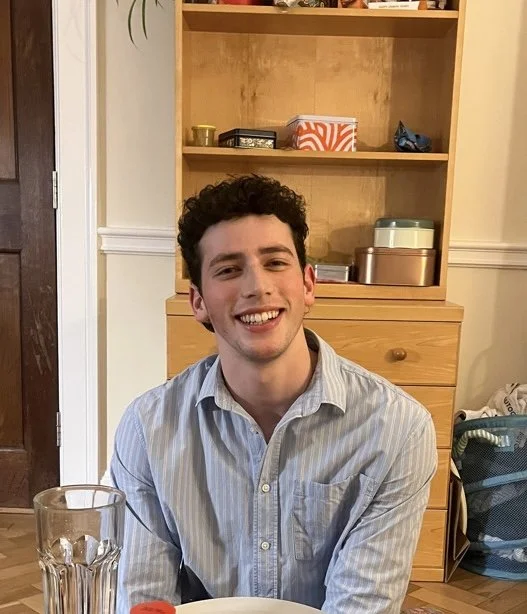 A young man with curly dark hair smiling and wearing a light blue striped shirt, sitting in a room with wooden furniture and a glass of water in front of him.