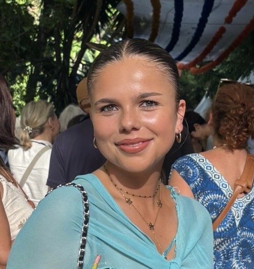 A young woman with light brown hair in a ponytail, wearing a light blue top, smiling at the camera during an outdoor event with other people and red, white, and blue decorations.