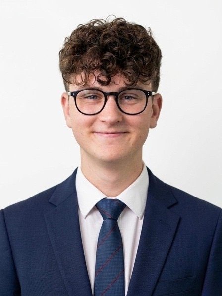 Headshot of a young man with curly brown hair, glasses, wearing a dark blue suit, white shirt, and a blue tie, smiling against a plain white background.
