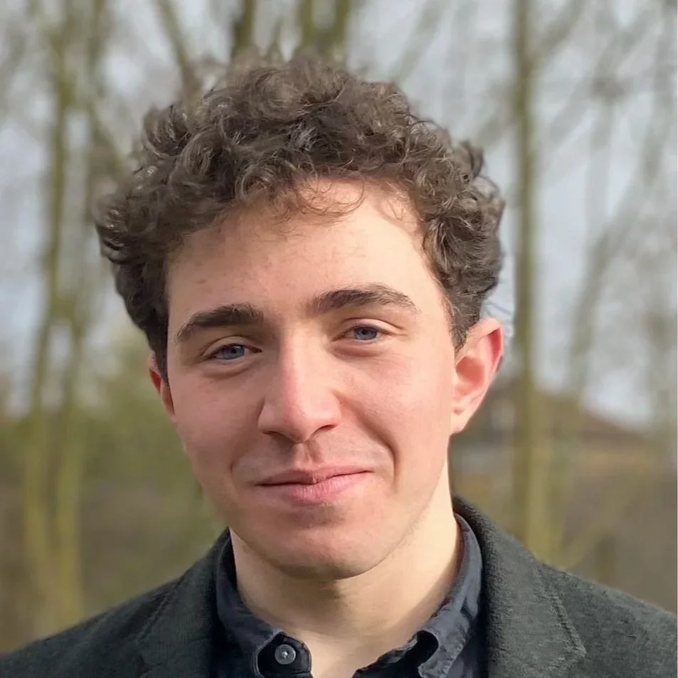 A young man with curly brown hair and blue eyes, smiling outdoors with trees in the background.