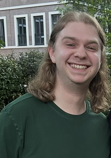 Smiling young man with long blonde hair wearing a green shirt outdoors in front of a building and greenery.