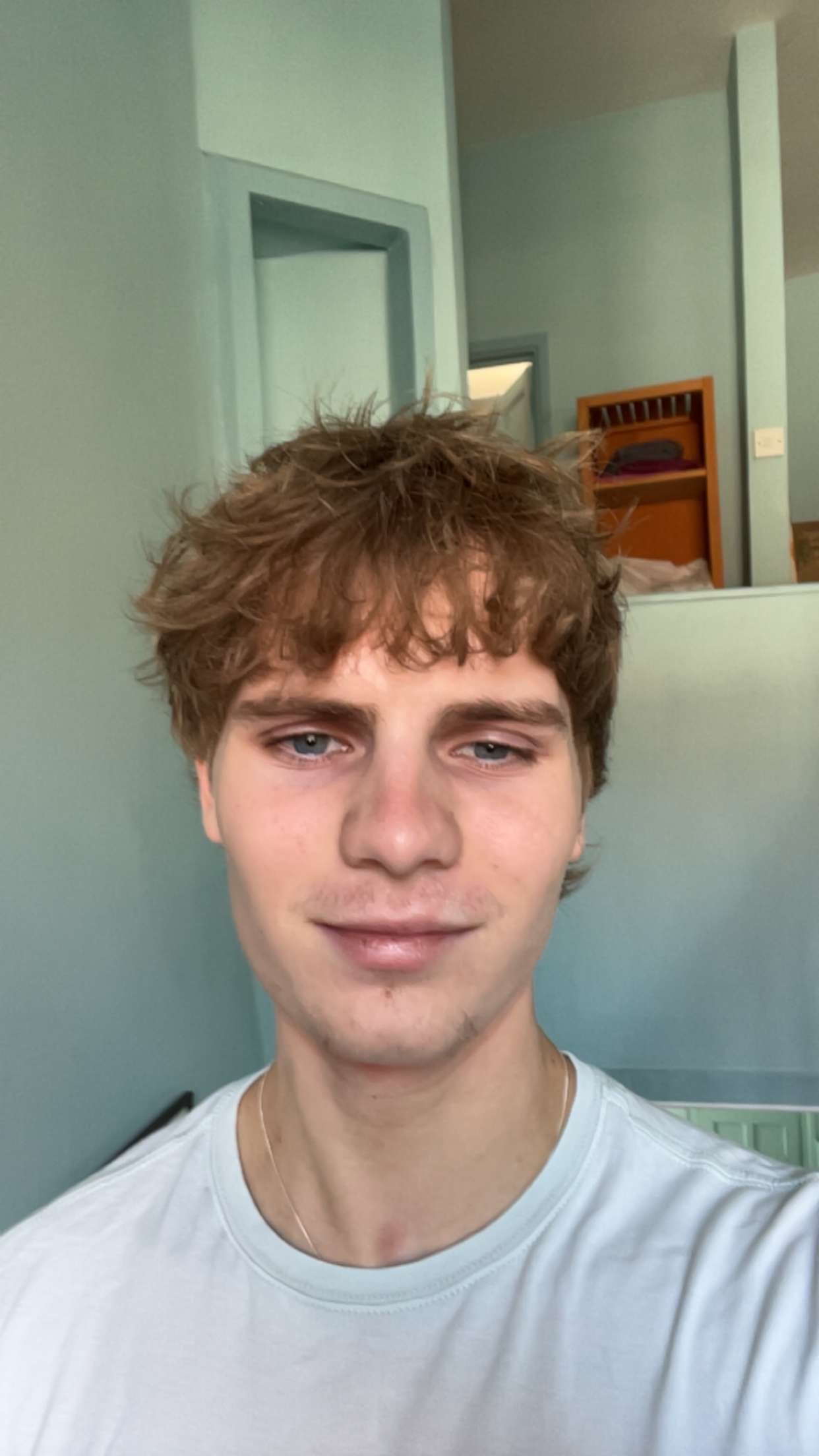 A young man with curly brown hair and blue eyes taking a selfie indoors, with a light green wall and a mirror in the background.