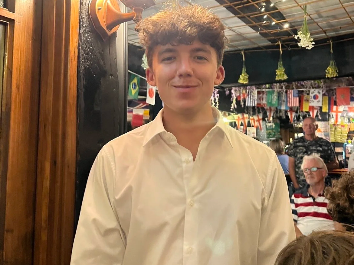 A young man with curly brown hair and a white button-up shirt standing in a restaurant or bar with international flags hanging in the background, and other people seated at tables.