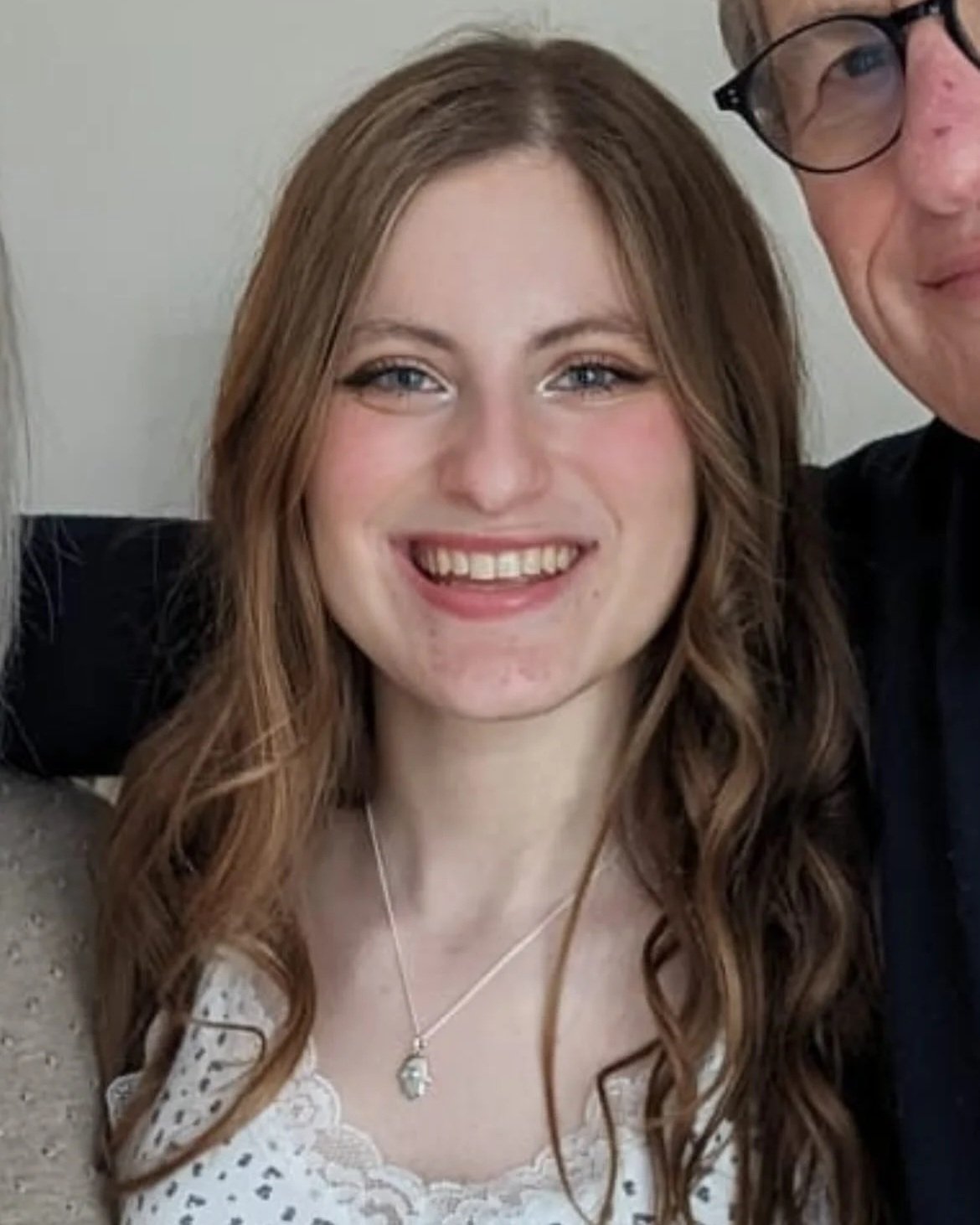 A young woman with long, wavy, auburn hair and blue eyes smiling at the camera. She wears a white lace top and a silver necklace with a pendant.