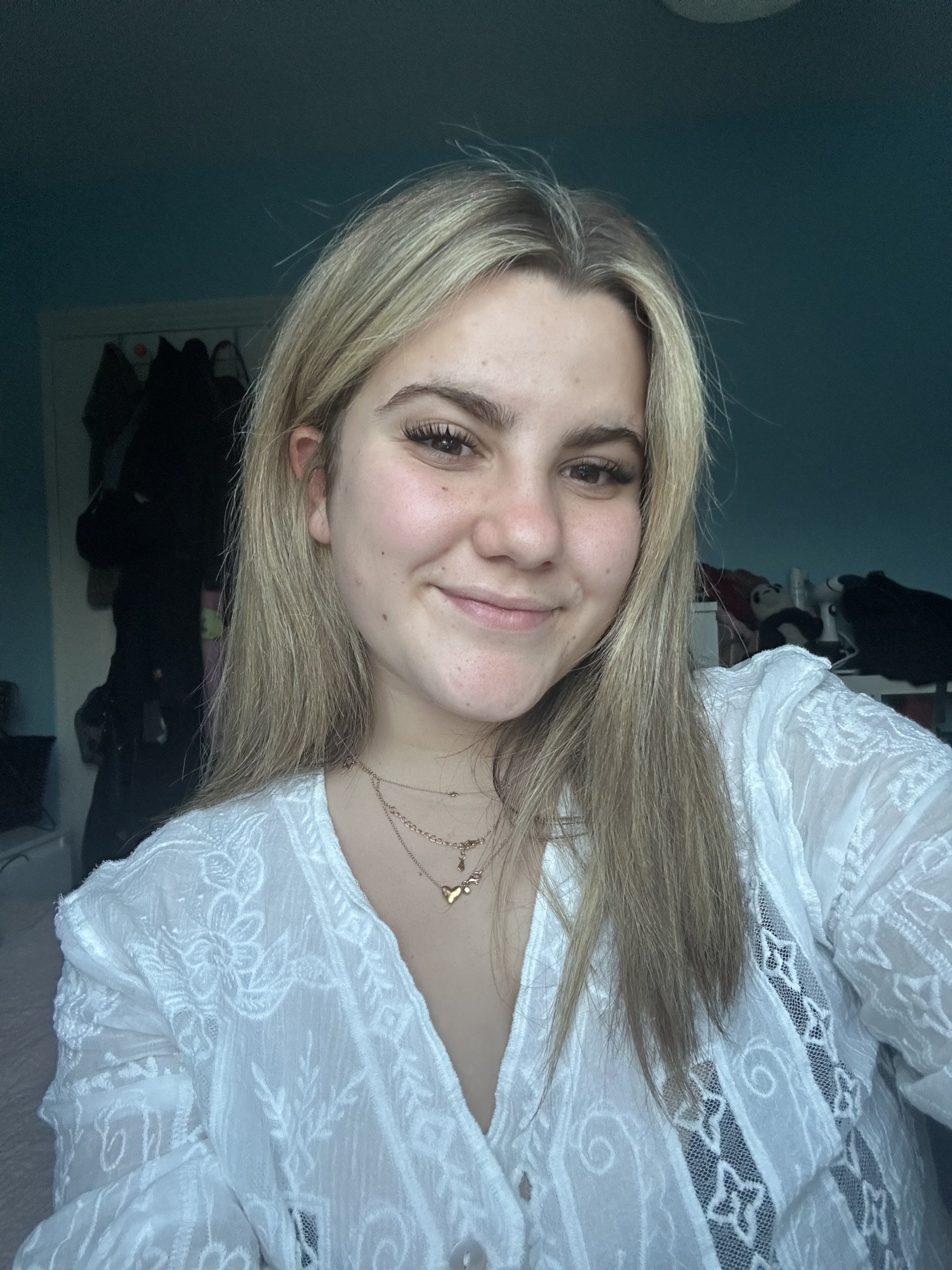 Close-up of a young woman with blonde hair smiling, wearing a white embroidered dress and layered gold necklaces, in a room with teal walls and a dresser with various items in the background.