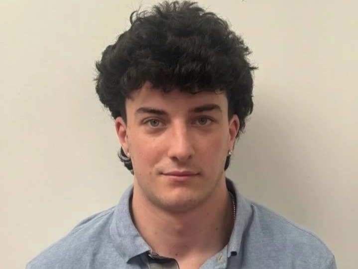 A young man with fair skin and dark, curly hair, wearing a gray collared shirt, standing against a light-colored wall.