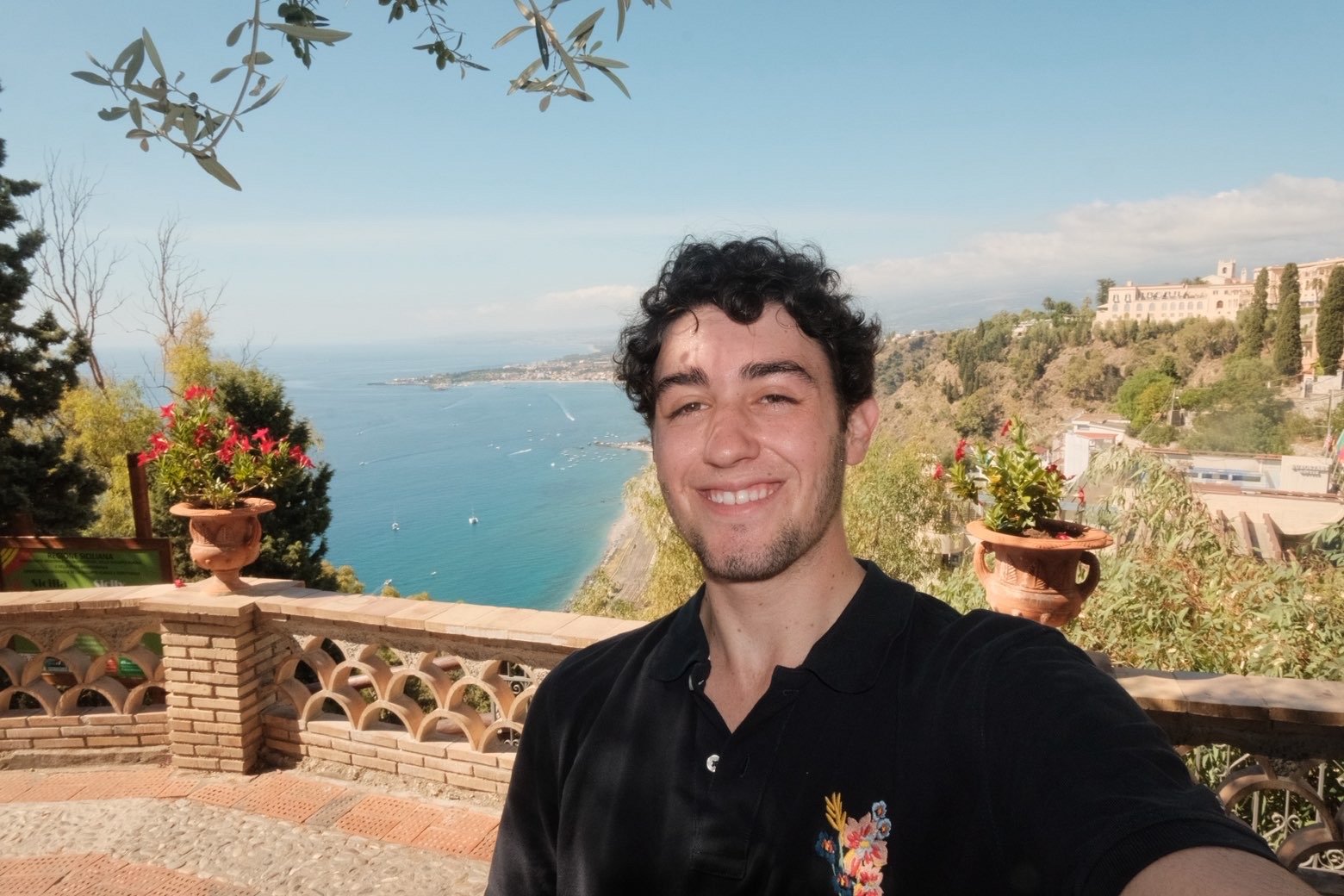 A young man taking a selfie outdoors with a scenic coastal view in the background. The scene includes blue water, boats, greenery, and buildings on a hillside, with a clear sky above.