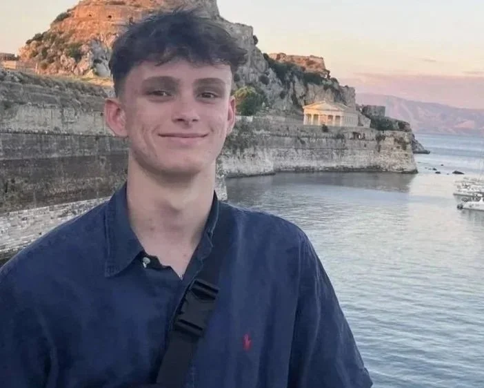 A young man with short dark hair, wearing a dark blue button-up shirt and a black strap across his chest, standing in front of a body of water with cliffs and a historical structure in the background during sunset.