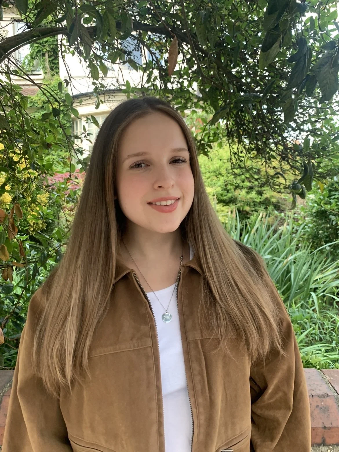 A young woman with long, light brown hair, wearing a tan jacket and a white shirt, standing outdoors surrounded by lush green foliage.