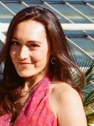 A young woman with long brown hair, smiling, wearing a pink scarf or top, and standing outdoors near water with plants and a bridge in the background.