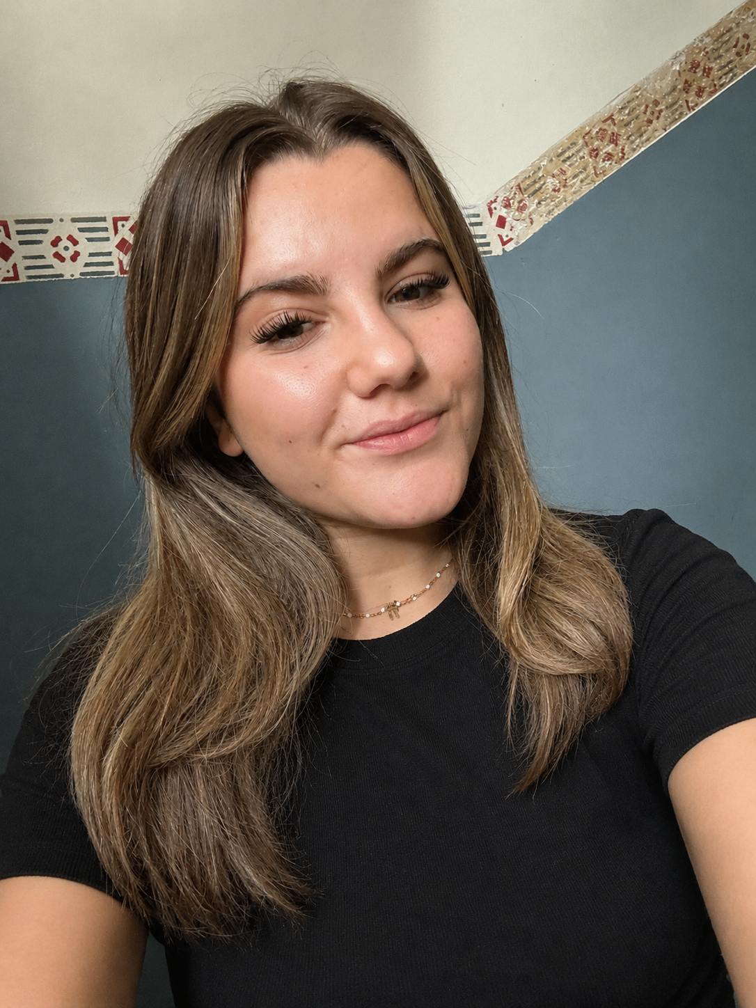 A young woman with long, wavy brown hair taking a selfie indoors. She is wearing a black shirt and a delicate necklace. The background features a two-tone wall with a patterned border.