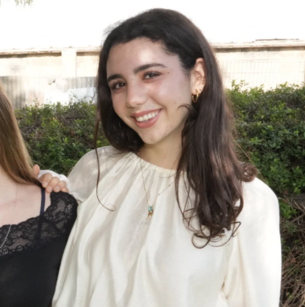 A young woman with long dark hair, gold hoop earrings, and a gold necklace with a turquoise pendant, smiling outdoors, wearing a cream-colored blouse.