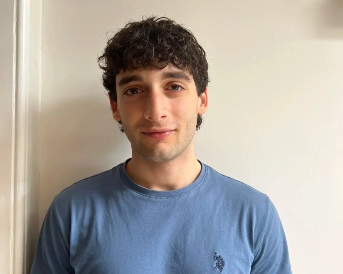 A young man with curly dark hair wearing a blue t-shirt, standing in front of a plain off-white wall.