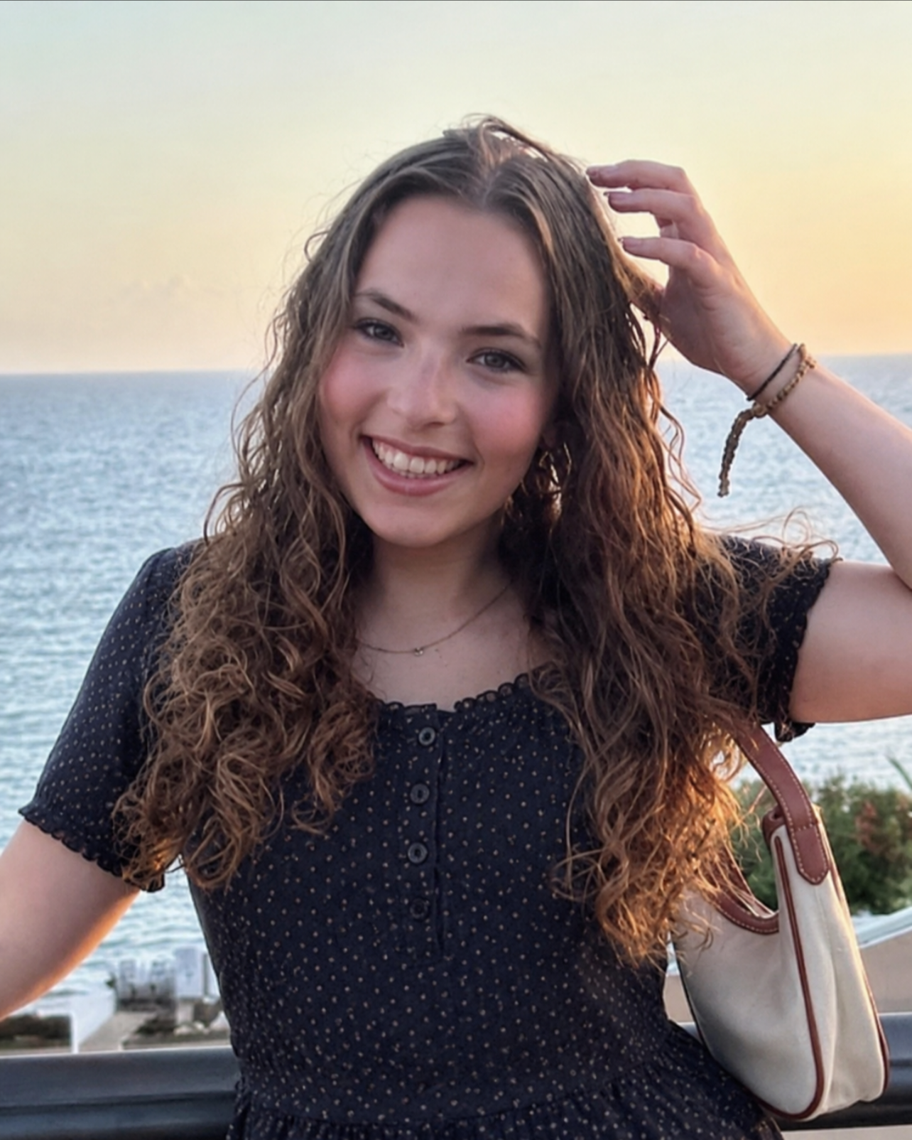 A young woman with long curly hair smiling at the camera, wearing a navy blue polka dot dress, standing outdoors near a body of water at sunset.