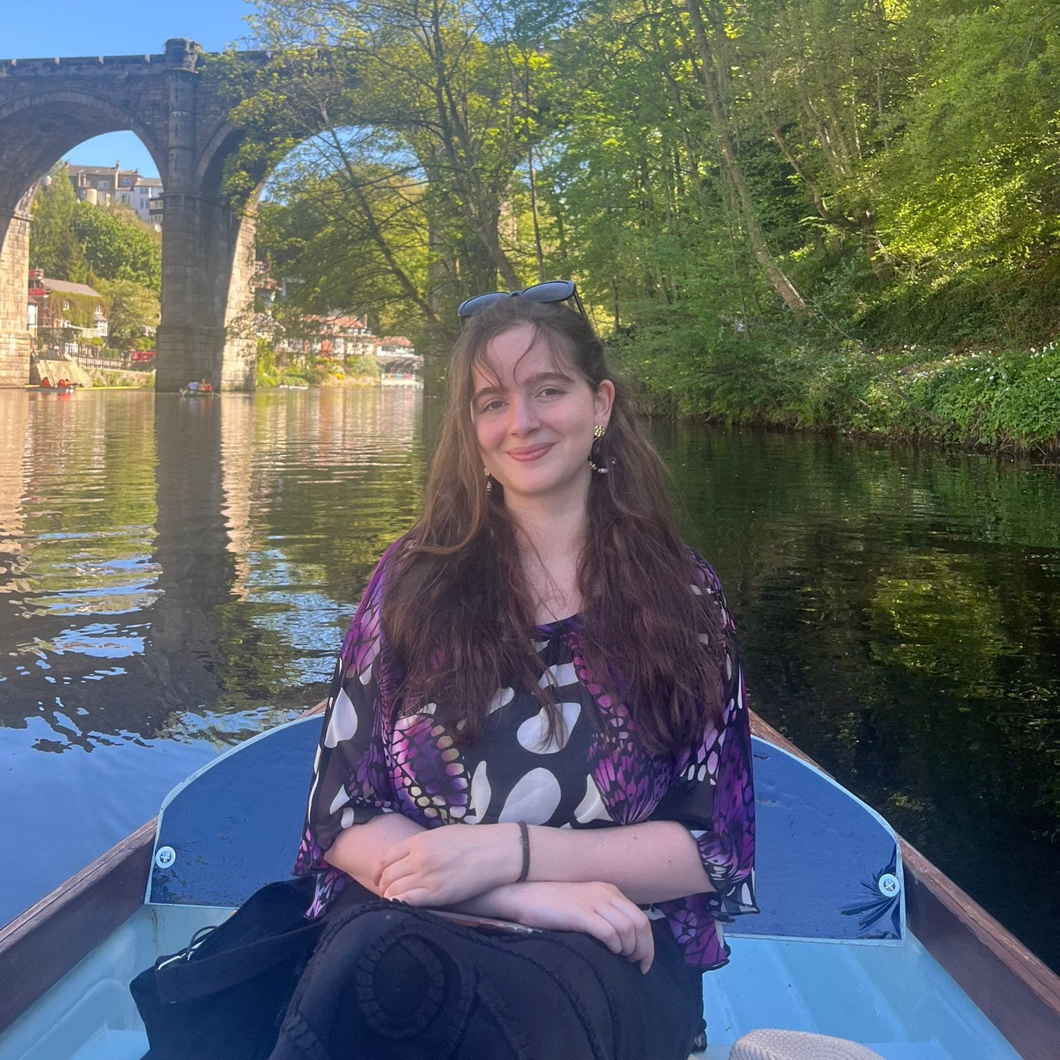 A woman with long brown hair, wearing a colorful patterned blouse, sitting in a boat on a river, with a stone arch bridge and green trees in the background on a sunny day.