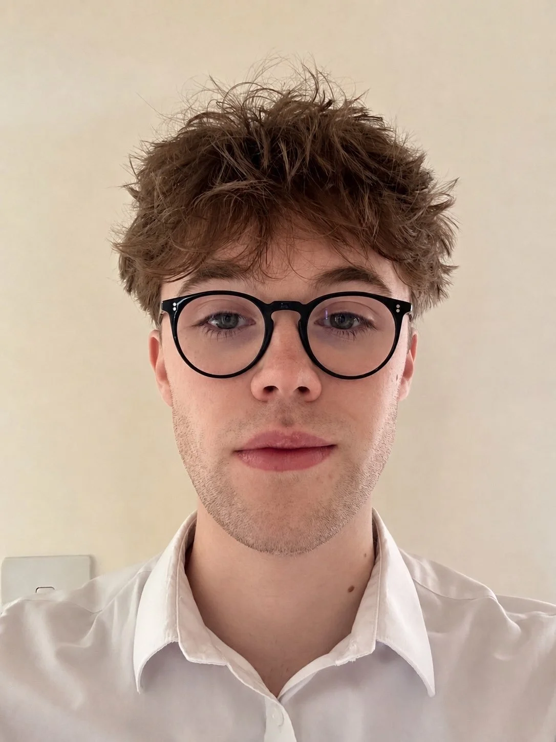 A young man with curly brown hair, wearing round black glasses and a white dress shirt, looking directly at the camera against a plain background.