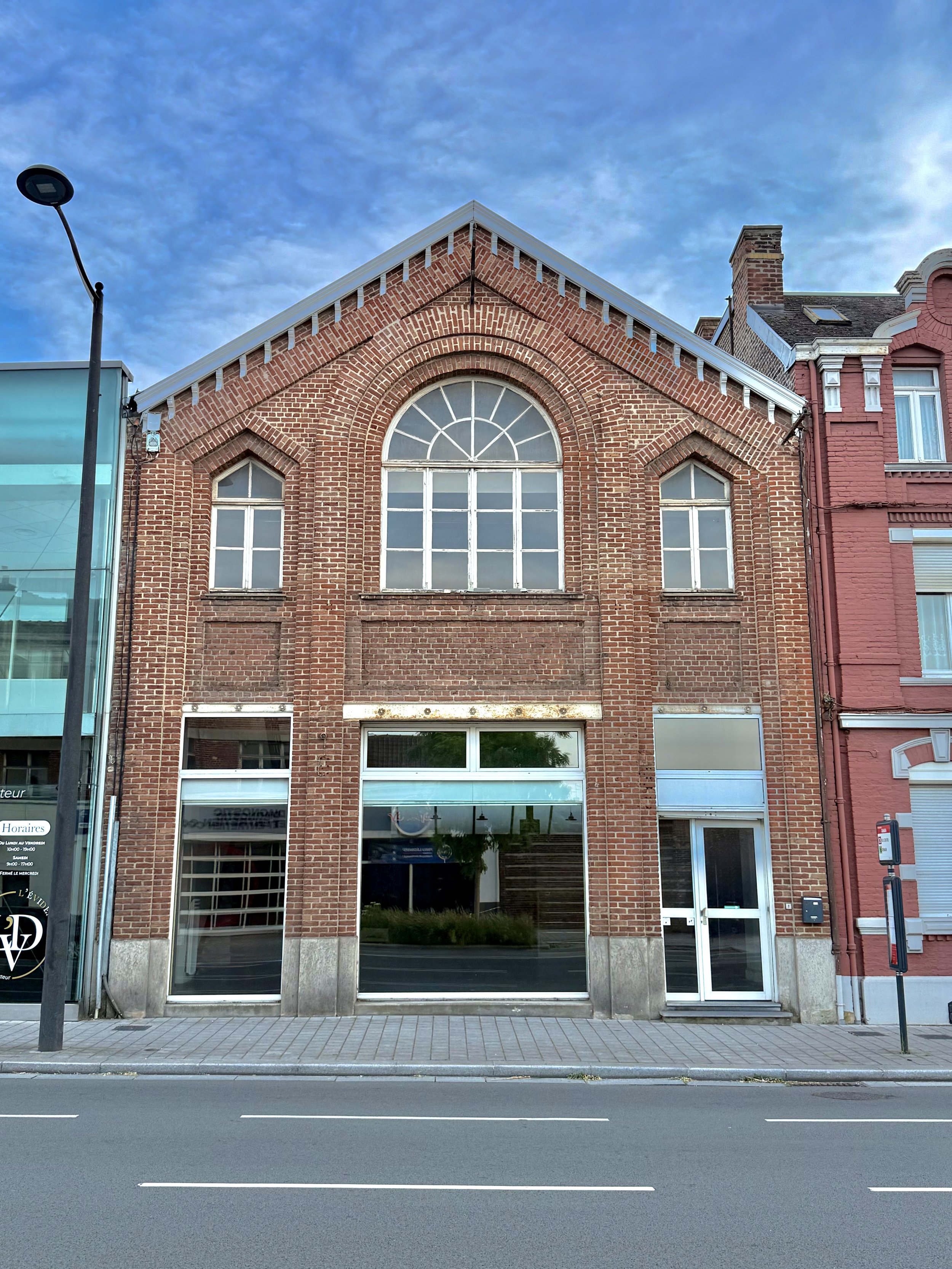 Un bâtiment en briques rouges avec de grandes fenêtres, situé dans une rue avec un trottoir et une route. Le bâtiment a un toit en pente et des fenêtres arrondies. Il y a une lampe de rue et une affiche publicitaire à gauche, et une boîte aux lettres à droite.