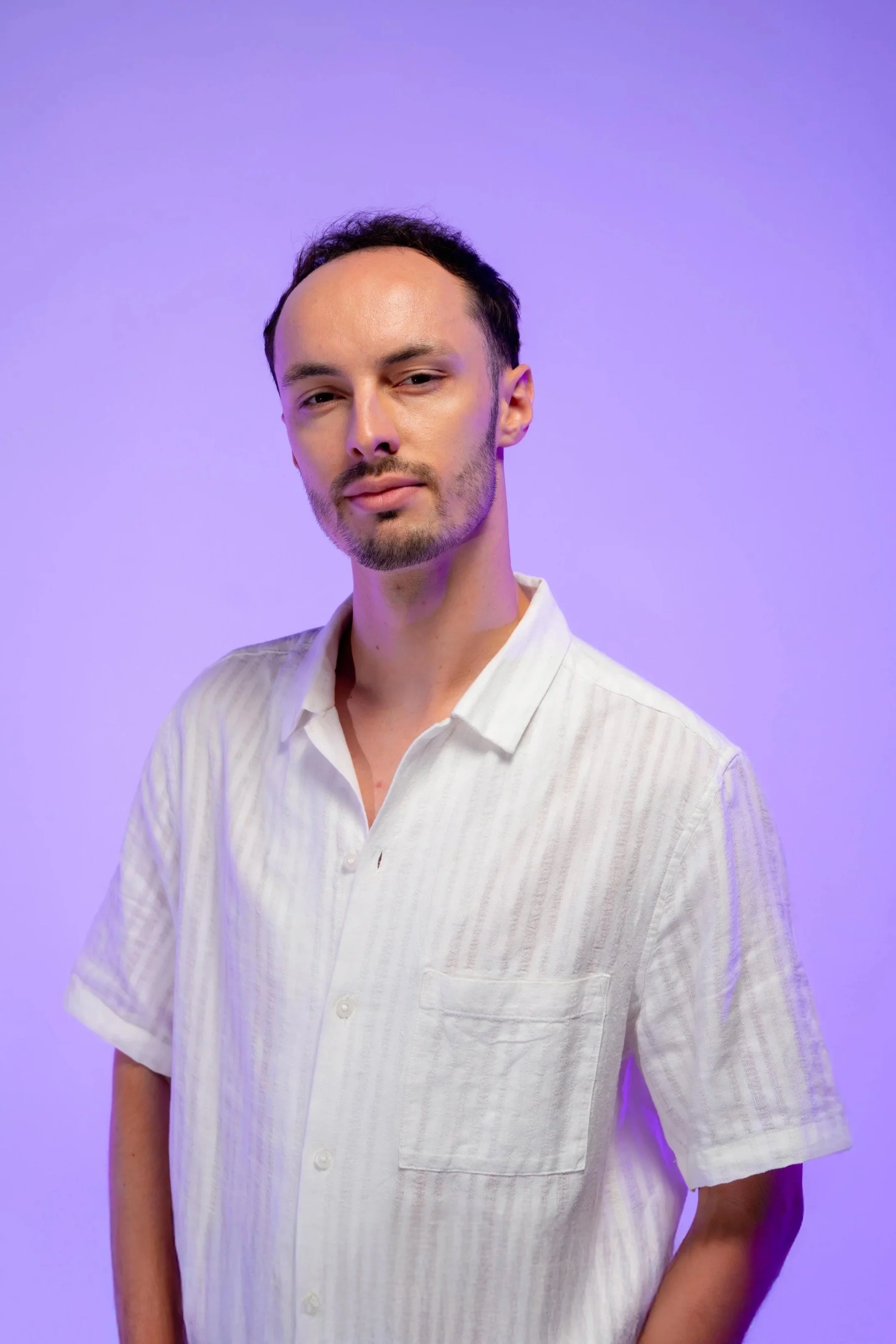 Portrait d'un homme avec une peau claire, portant une chemise blanche à rayures, se tenant contre un fond violet avec un éclairage doux.