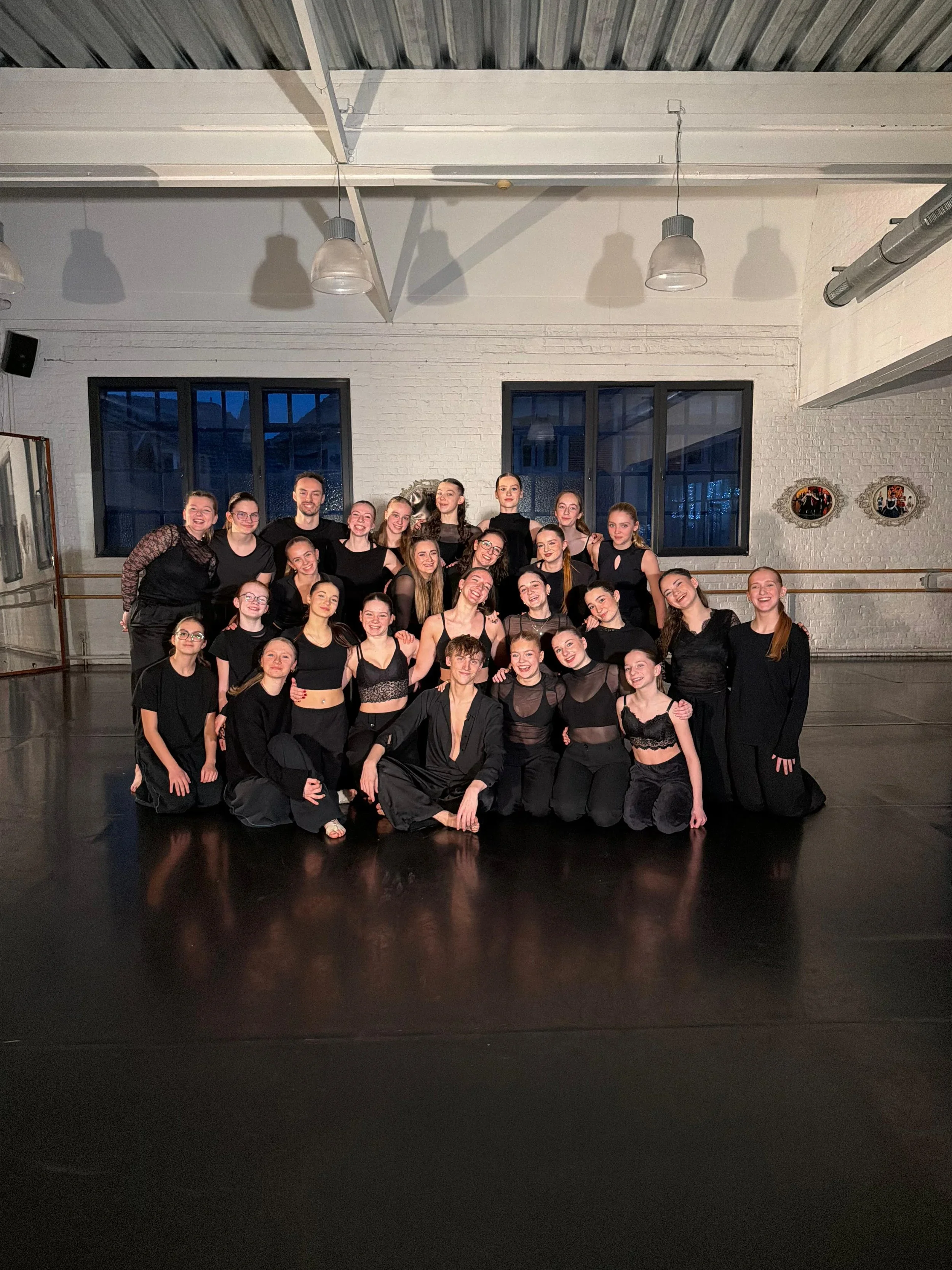 Groupe de personnes posant pour une photo dans une salle de danse ou de studio, certaines en vêtements de danse noirs, souriantes et heureuses.