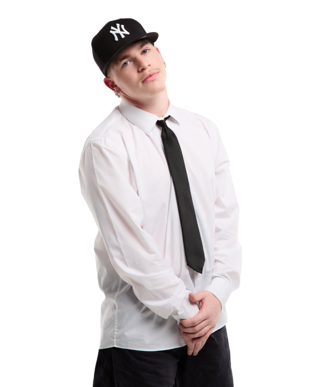 Jeune homme portant une casquette New York, une chemise blanche, une cravate noire, et des boucles d'oreilles, posant contre un fond blanc.